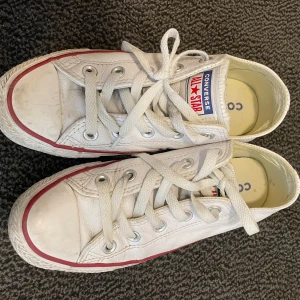 W Converse All Star sneakers - Säljer ett par klassiska vita Converse All Star sneakers i storlek 36. Skorna har lågt skaft, snörning och är tillverkade i skinn med röd och blå rand längs sulan. 