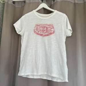 Vit t-shirt med röd print Authentic - Vit t-shirt i bomull med korta ärmar och rund hals. Framsidan har ett rött, lite slitet tryck med texten 'AUTHENTIC' i retrostil. Trycke är tyckt på insidan. De fins en liten fläck på magen som knappast syns.