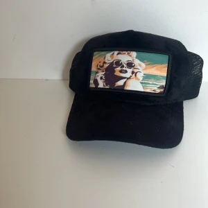 Svart keps från Revenite med tryck - Svart keps från Revenite med mesh på sidorna och justerbar snapback. Framtill finns en färgglad patch med retroinspirerat motiv av en kvinna vid havet. Kepsen levereras med en svart påse med Revenite-logga. Perfekt för dig som vill sticka ut.