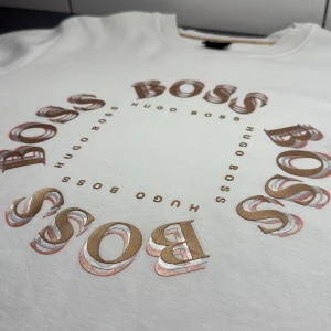 Vit sweatshirt från Hugo Boss - Vit sweatshirt från Hugo Boss med stort cirkelformat BOSS-tryck i guld, silver och beige på bröstet. Tröjan har rund halsringning och långa ärmar. Tillverkad i mjuk bomull för en skön och avslappnad stil. Använd 1 gång, den var för stor på mig! Mycket bra Skick 