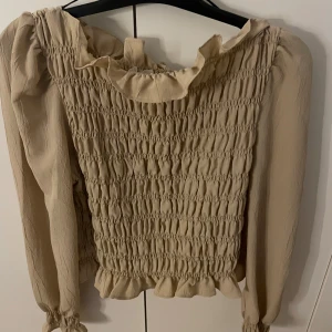 Beige blus med volang och smock - Säljer en beige blus med smockad front och volangdetaljer vid hals och nederkant. Blusen har långa ärmar med knyt vid ärmslut och en mjuk, luftig känsla. Perfekt för dig som gillar romantiska och trendiga detaljer.