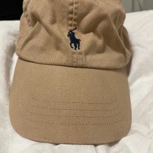 Beige keps från Polo Ralph Lauren - Snygg beige keps från Polo Ralph Lauren med klassisk blå broderad logga framtill. Nypris 700kr säljer då ja köpte fel färg 