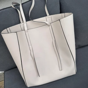 Vit shoppingväska i skinn - Stor och stilren shoppingväska i ljusvit färg med raka linjer och tunna handtag. Väskan har minimalistisk design och är tillverkad i slätt skinnimitation med svarta kanter som kontrast. Perfekt för dig som gillar enkel och clean look. Ljusgrå / beige toteväska. Ej slitage på handtag men Fläckig, var god se bilder. Jag har massor av artiklar till salu! 