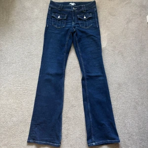 Blå lågmidjade bootcut jeans från Gina Tricot - Snygga mörkblå lågmidjade jeans från Gina Tricot i bootcut-modell. Klassisk femficksdesign med två framfickor med lock och knappdetalj, samt två bakfickor. Jeansen har kontrastsömmar och är tillverkade i jeansmaterial med skön stretch. Perfekt för dig som gillar retrovibbar och markerad siluett. Byxorna är i storlek 164 men passar även dem som oftast handlar på den vanliga kvinno avdelningen.