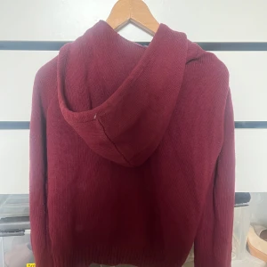 Vinröd stickad hoodie med dragkedja - Mysig vinröd hoodie i stickat material med huva och snörning. Tröjan har praktisk dragkedja framtill och två fickor. Perfekt för chill dagar och när du vill ha något bekvämt och snyggt. Ribbad kant vid ärmslut och nederkant för extra stil.