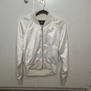 Vit satin bomberjacka från BikBok - Snygg vit bomberjacka från BikBok i satinliknande material. Jackan har dragkedja framtill, ribbade muddar och klassisk bomberkrage. Perfekt för dig som vill ha en clean och stilren look med lite glansig finish.