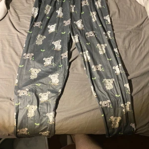 Grå mjukisbyxor med koalamönster - Supermysiga grå mjukisbyxor med gulligt koalamönster och små gröna blad. Perfekta för chill hemma eller som pyjamasbyxor. Byxorna har en avslappnad passform och är gjorda i mjukt material som känns skönt mot huden. Rekommenderas för barn mellan 10-13 år beronende på om man har långa ben eller ej