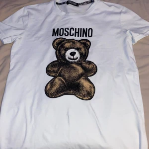 Vit Moschino t-shirt med björntryck - Snygg vit t-shirt från Moschino med stort tryck av en brun nalle på framsidan och Moschino-logga ovanför. Klassisk rund hals och korta ärmar. Perfekt statement-plagg för dig som gillar coola designer och streetstyle.
