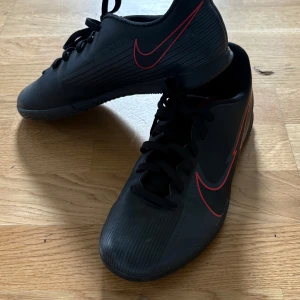 Nike Mercurial fotbollsskor svart/röd - Svarta Nike Mercurial fotbollsskor med röda detaljer och klassisk Swoosh-logga på sidan. Skorna har snörning och en slitstark syntetsula med grepp, perfekt för inomhusspel. Snygg, sportig design som sticker ut på planen.