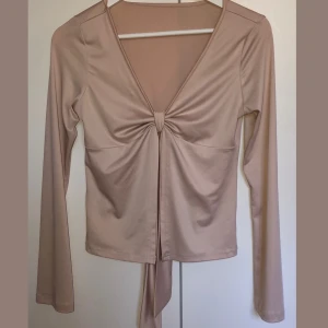 Beige långärmad omlottopp med knyt - Trendig beige topp med lång ärm och djup v-ringning. Snygg knytdetalj framtill som ger en omlottkänsla. Mjuk och stretchig i syntetmaterial, perfekt för att skapa en clean och stilren look.