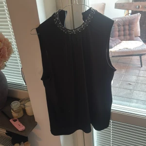 Svart pärlbroderad ärmlös topp Stockh LM - Supersnygg svart ärmlös topp från Stockh LM med hög hals och detaljerat pärl- och paljettbroderi runt kragen. Toppen har en rak passform och stängs med dragkedja i nacken. Perfekt för dig som vill sticka ut med lite extra bling. Tycker att denna topp är jättefin men den passar inte mig så som jag vill, jag rekommenderar starkt!