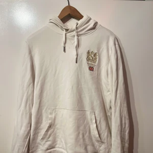 Vit hoodie från Morris med tryck - Säljer en vit hoodie från Morris med snyggt broderat emblem och brittisk flagga på bröstet. Tröjan har huva med snörning, magficka och långa ärmar. Perfekt för chill dagar och enkel att matcha med jeans eller joggers.