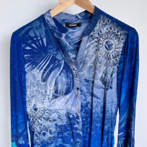 Blå långärmad skjorta från Desigual - Snygg blå skjorta från Desigual med lång ärm och knappar framtill. Skjortan har ett unikt mönster med vita och blå detaljer som ger en cool och konstnärlig vibe. Materialet känns lätt och bekvämt, perfekt för dig som gillar färgstarka och kreativa plagg.