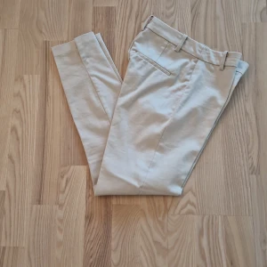 Beige kostymbyxor från H&M, stl 32 - Oanvända snygga beige kostymbyxor från H&M i storlek 32. Byxorna har raka ben, bälteshällor och diskreta fickor bak. Perfekta för en clean och stilren look. Tillverkade i mjukt bomullsmaterial som känns skönt mot huden.