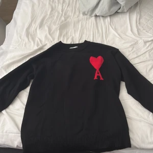 Svart stickad tröja från Ami Paris - Svart stickad tröja från Ami Paris med lång ärm och ribbade muddar. På bröstet finns ett stort rött hjärta med bokstaven A, som ger en unik och trendig vibe. Perfekt för dig som gillar stilrena plagg med en twist.