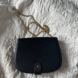 Svart axelväska med guldkedja - Stilren svart axelväska i skinn med guldig kedja. Den går att ha med långt band eller vika bandet och ha över axeln. Lappen är borta så vet inte vart väskan är ifrån.