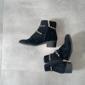 Svarta boots med kedjedetaljer River Island - Snygga svarta boots från River Island i mocka med chunky klack och rund tå. Dekorerade med guldfärgade kedjor och dragkedja på sidan för enkel på- och avtagning. Perfekt för dig som gillar att sticka ut med coola detaljer och stilren look.