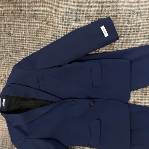 Marinblå kostym från Calvin Klein - Snygg tvådelad marinblå kostym från Calvin Klein med klassisk kavaj och matchande kostymbyxor. Kavajen har två knappar framtill, fickor med lock och stilren passform. Materialet är en blandning av polyester och rayon med svart foder. Perfekt för dig som vill ha en clean och modern look. Använt ett fåtal gånger och är pojk storlek 10