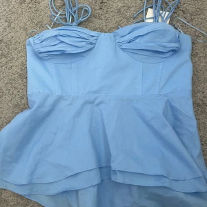 Ljusblå peplum linnetopp med snörning - Supersöt ljusblå linnetopp med peplumvolang och smala axelband med snörning. Toppen har rynkade kupor och smock i ryggen för extra komfort. Perfekt för en somrig look och lätt att matcha med jeans eller kjol.