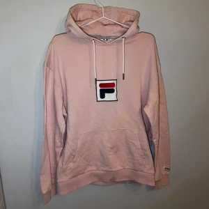 Rosa hoodie från FILA med logga - Säljer en chill rosa hoodie från FILA i storlek M. Tröjan har stor huva med snörning, magficka och den klassiska FILA-loggan broderad på bröstet. Perfekt för att slänga på sig när det är lite kyligt ute.