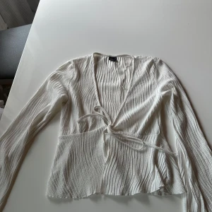 Vit omlottopp från Gina Tricot - Supersnygg vit omlottopp från Gina Tricot med ribbad struktur och långa ärmar. Toppen har knyt i sidan och djup v-ringning, vilket ger en trendig och fräsch look. Perfekt att styla med jeans eller kjol för en cool vibe.