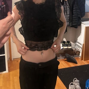 Svart spetsig blus  - Säljer en svart croppad blus med detaljerad spets och blommigt mönster. Toppen har ärmlös design med volangkant vid axlarna och hög hals. Baksidan är transparent med dragkedja. Perfekt för dig som gillar statement, plagg med mycket detaljer. Storlek s men xs. Knappt använd. 