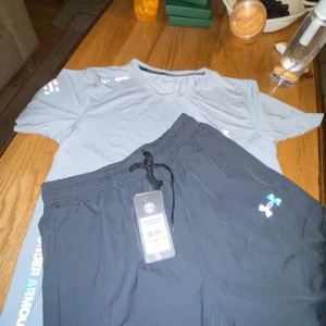 Grå Under Armour t-shirt för träning - Snygg grå t-shirt från Under Armour med korta ärmar och coolt holografiskt tryck på bröstet och axeln. Perfekt för dig som gillar sportig stil och vill ha något som andas bra under träningen. Materialet är lätt och känns skönt mot huden.