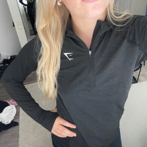 Gymshark tröja - Svart långärmad tröja från Gymshark med half zip och tumhål i ärmsluten. Snyggt sportigt snitt och diskret logga på bröstet. Perfekt för träning eller chill dagar. Materialet är mjukt och stretchigt, troligen polyester eller syntet.
