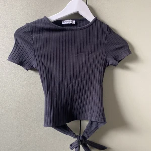 Svart ribbad knyt-topp - Svart ribbad topp med korta ärmar. Toppen har snygg knytning baktill som ger en cool detalj. Tillverkad i mjuk bomullsmix som känns skön mot huden. Perfekt till jeans eller kjol för en trendig look. Pris kan förhandlas.