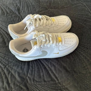 Nike Air Force 1 vit/guld sneakers - Nike Air Force 1 sneakers i vit med snygga gulddetaljer på logga, snörning och lace tag. Klassisk låg modell med perforerad tå och platt sula. Ovandel i skinn och meshfoder för extra komfort. Perfekt för dig som vill ha en clean och stilren look med lite extra bling.