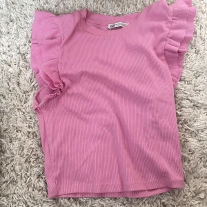 Rosa croppad topp med volang från Zara - Supersöt rosa croppad topp från Zara med ribbad struktur och volangdetaljer på axlarna. Toppen är ärmlös och har en rund halsringning. Perfekt att styla med jeans eller kjol för en trendig look.💗💗