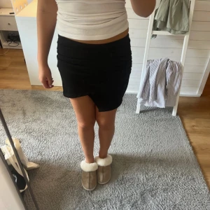 Svart omlottkjol med slits - Snygg svart kortkjol med omlott-design och diskret slits framtill. Kjolen har en tight passform och är enkel att matcha med olika toppar. Perfekt för dig som gillar stilrena och trendiga plagg.