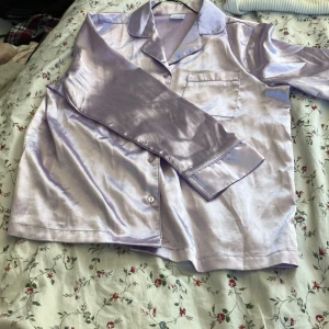 Ljuslila satinpyjamas skjorta - Snygg ljuslila skjorta i satin-look med klassisk krage, bröstficka och knappar framtill. Skjortan har långärmade ärmar och glansigt material som ger en lyxig vibe. Perfekt för chill hemma eller sleepover med kompisarna.