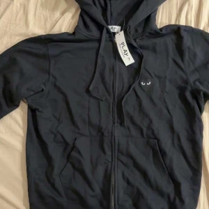 Svart hoodie från Comme des Garçons Play - Svart hoodie från Comme des Garçons Play med dragkedja, huva och snörning. Ikoniskt hjärta med ögon broderat på bröstet. Mjuk bomullskvalitet och praktiska fickor framtill. Perfekt för dig som gillar stilrena streetwear-plagg.