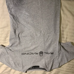 Grå långärmad tröja Sail Racing - Snygg grå långärmad tröja från Sail Racing med svart logotyp tryckt på bröstet och nertill. Tröjan har rund hals och är tillverkad i mjuk bomull, perfekt för chill dagar eller när du vill ha en clean look. Passar dig som gillar sportiga och stilrena plagg.