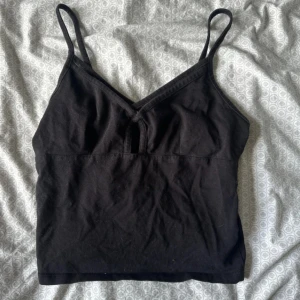 Svart linnetopp från Gina Tricot - En svart linnetopp från Gina Tricot med smala axelband och v-ringning framtill. Toppen är croppad och har en liten öppning vid bysten för extra detalj. Perfekt att matcha med jeans eller kjol för en enkel och snygg look.