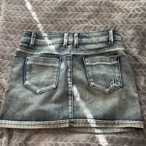 Blå jeanskjol från d.brand - Snygg lågmidjad jeanskjol från d.brand i klassisk blå tvätt med slitningar och kontrastsömmar. Kjolen har fem fickor, bälteshällor och dragkedja med knapp framtill. Bakfickorna har broderad logga och cool faded effekt. Storlek 26