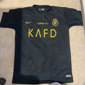 Al Nassr Ronaldo 7 Nike fotbollströja - Snygg mörkblå Al Nassr fotbollströja från Nike med gult tryck, inklusive klubbmärke, sponsorloggor och Ronaldo 7 på ryggen. Tröjan är kortärmad och tillverkad i ett lätt, andningsbart material perfekt för match eller träning.