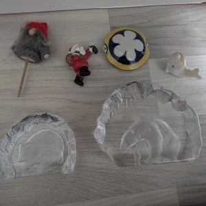 Olika prydnadsföremål i paket - Bilden visar flera prydnadsföremål, bland annat två glasfigurer med djurmotiv (igelkott och björn), en tomte, en liggande jultomte, en liten val och ett runt föremål med blomdekor. Ingen bok syns på bilden.
