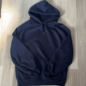 Mörkblå hoodie med huva - Säljer en enkel och stilren mörkblå hoodie med huva. Tröjan har långa ärmar, ribbade muddar och en klassisk loose passform. Perfekt för chill dagar eller när du vill ha något bekvämt och snyggt. Materialet känns mjukt och är troligtvis bomull eller polyester.