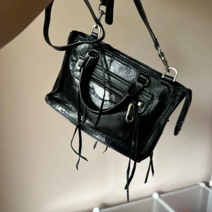 Svart handväska från Rebecca Minkoff - Säljer en svart handväska från Rebecca Minkoff i skinn med både handtag och avtagbar axelrem. Väskan har silverfärgade metalldetaljer, dragkedja framtill och dekorativa skinnband. Kommer med original dustbag.