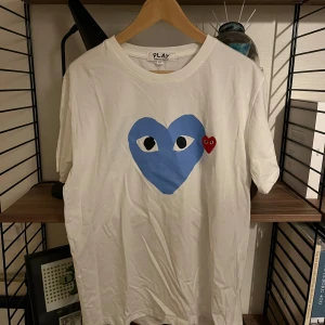 Vit Comme des Garçons Play t-shirt 2XL - Snygg vit t-shirt från Comme des Garçons Play i storlek 2XL. Sitter dock mera som en L/XL. Framsidan har ett stort blått hjärta med ögon och ett mindre rött hjärta bredvid. Klassisk rund hals och korta ärmar. Materialet är mjuk bomull, perfekt för en chill och trendig look.
