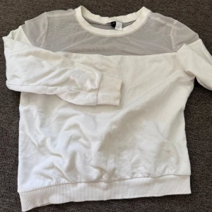 Vit tröja med mesh  - Säljer en vit långärmad tröja från DIVIDED i storlek M. Tröjan har en snygg meshdetalj över axlar och bröst, vilket ger en cool kontrast till det mjuka bomullstyget. Rund hals och ribbade muddar vid ärmslut och nederkant. Perfekt för dig som gillar stilrena plagg med en twist.