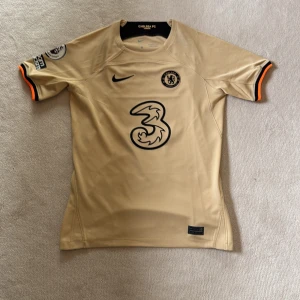 Chelsea FC beige matchtröja Nike - Chelsea FC fotbollströja i beige med svarta detaljer och orange kant på ärmarna. Nike-logga på bröstet, klubbmärke och stor sponsorlogga i svart. Tillverkad i lätt, ventilerande Dri-FIT-material som passar perfekt på planen. Snygg och modern design för fansen. Den är aldrig använd.