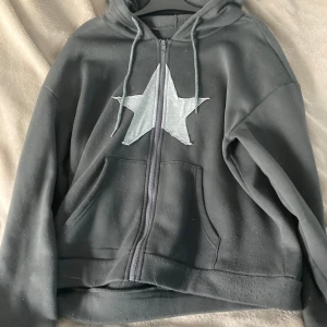 Grå/blå hoodie med grå stjärna - Grå/blå hoodie med dragkedja och huva, prydd med en stor grå stjärna på bröstet. Tröjan har långa ärmar, snörning vid huvan och praktiska fickor framtill. Perfekt för chill dagar och enkel att matcha med jeans eller mjukisbyxor.