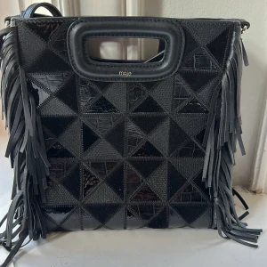 Svart axelväska med fransar från Maje - Säljer en svart axelväska från Maje med coola fransar på sidorna och geometriskt mönstrad framsida i olika texturer. Väskan har inbyggda handtag och axelrem, tillverkad i skinn. Perfekt för dig som vill sticka ut med en unik accessoar. Aldrig använd, och köpt på NK 