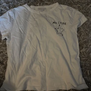 Vit t-shirt med stjärntryck Gina Tricot - Vit t-shirt från Gina Tricot med svart tryck av texten 'MA CHÉRIE' och en stjärna både fram och bak. Klassisk rund hals och korta ärmar. Skön och enkel modell i mjuk bomull, perfekt till jeans eller shorts.