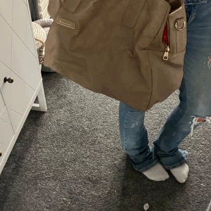 beige väska  - Stor beige axelväska från Björn Borg i skinnimitation med flera fack och dragkedjor. Insidan är fodrad i rött tyg med logga. Perfekt för dig som vill ha plats för allt och gillar stilren design med praktiska detaljer.