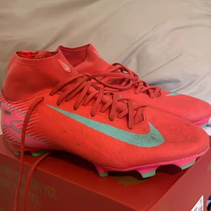 Röda Nike fotbollsskor med turkos swoosh - Säljer ett par riktigt snygga fotbollsskor från Nike i en stark röd färg med turkos swoosh och detaljer. Skorna har snörning, högre skaft och dobbar i grönt och rosa undertill. Perfekta för dig som vill sticka ut på planen.