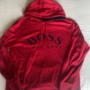 Röd hoodie från Hugo Boss med logga - Snygg röd hoodie från Hugo Boss i storlek S. Tröjan har en stor svart BOSS-logga på bröstet, huva med snörning och långa ärmar. Tillverkad i mjuk bomull som är skön mot huden. Perfekt för dig som gillar streetwear och vill sticka ut med en ikonisk look.